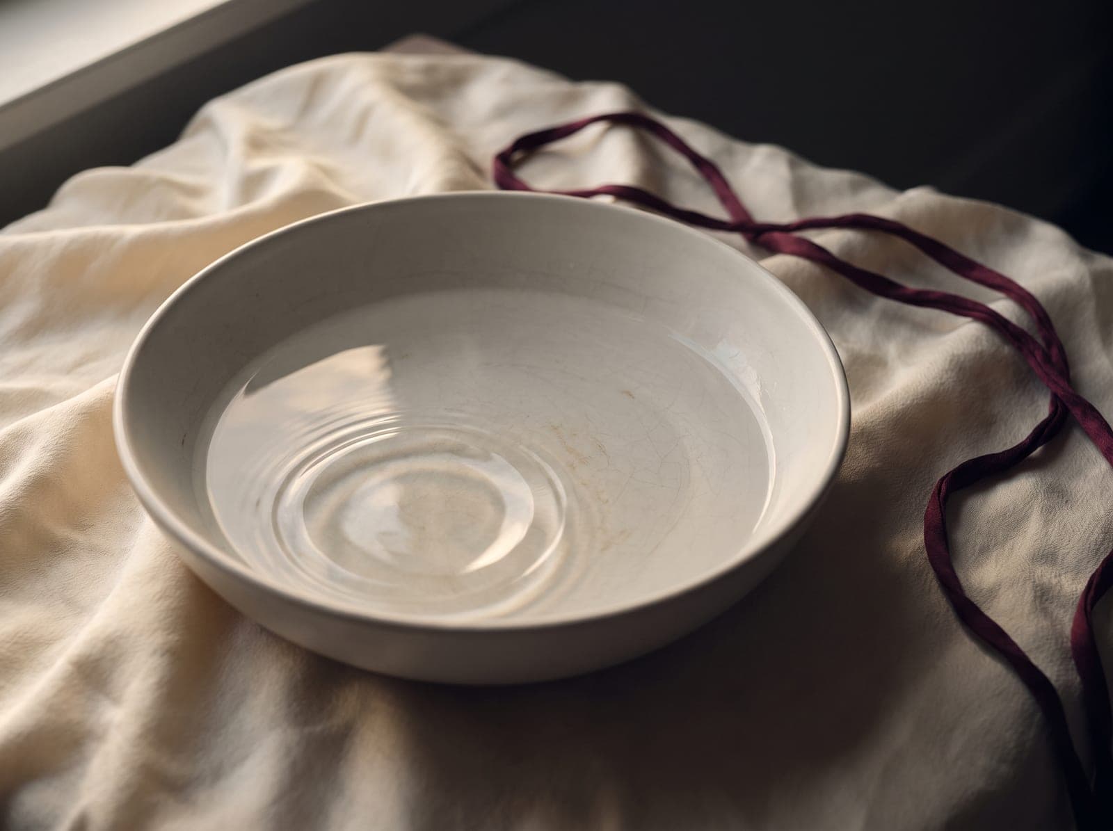 Still water with gentle ripples in a ceramic bowl on cream silk, lit by warm window light